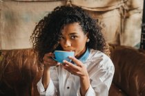 Lockige junge Frau trinkt eine Tasse Kaffee und blickt in die Kamera — Stockfoto