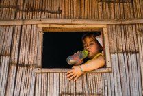 CHIANG RAI, THAILAND- FEVEREIRO 12, 2018: Menina sentada à janela na cabana e tomando leite de garrafa de plástico . — Fotografia de Stock