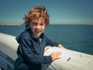 Joyeux jeune garçon appuyé sur la main courante blanche au bord de la mer et regardant la caméra . — Photo de stock