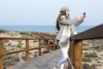 Cheerful woman taking shots with smartphone on boardwalk at seaside — Stock Photo