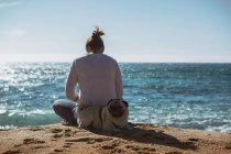 Вид сзади на женщину, сидящую на берегу моря с собакой-мопсом — стоковое фото