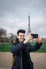Hombre alegre de pie y tomando selfie con teléfono inteligente sobre el fondo de la torre Eiffel . - foto de stock