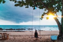 Back view of woman sitting on swings and looking at sunset at seaside. — Stock Photo