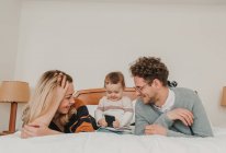Cheerful couple lying on bed with little kid playing with smartphone. — Stock Photo