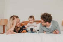 Веселая пара смотрит на ребенка со смартфоном на кровати — стоковое фото