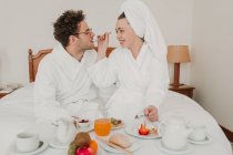Femme en peignoir nourrissant homme avec petit déjeuner dans le lit de l'hôtel — Photo de stock