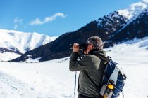 Vista lateral do fotógrafo sênior tirando fotos no prado nevado — Fotografia de Stock
