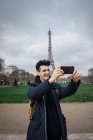 Joven en gafas de pie con el teléfono y tomando selfie en el fondo de la torre Eiffel . - foto de stock