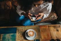 Desde arriba mujer sentada en la cafetería y tomando fotos de taza de café con leche con teléfono inteligente . - foto de stock
