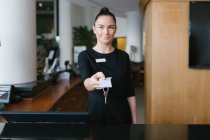 Hübsche brünette Hotelangestellte steht an der Rezeption und gibt Schlüssel. — Stockfoto