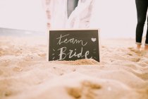 Доска с надписью невесты команды на песке и ногах урожая людей . — стоковое фото