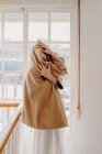 Side view of woman in stylish coat embracing herself at window. — Stock Photo