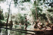 LAOS, LUANG PRABANG: Hombre local caminando con botella y balanceándose en el tronco sobre el estanque en el bosque soleado . - foto de stock