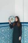 Молода красива жінка позує з барвистою тарілкою на фоні синьої плиткової стіни . — стокове фото