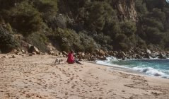 Зріла жінка в супроводі мопса собаки читає книгу на березі моря в сонячний день — стокове фото
