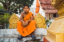 LAOS- FEBRERO 18, 2018: Niño monje sentado en la cerca y mirando a la cámara - foto de stock