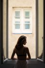 Вид сзади девушки, позирующей у окна дома и смотрящей через плечо в камеру — стоковое фото