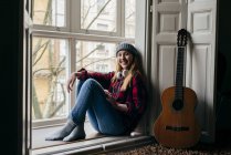 Lächelnde junge Blondine sitzt auf Fensterbank bei der Gitarre und blickt in die Kamera. — Stockfoto