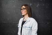 Mujer joven en gafas de pie en la pared oscura y mirando hacia otro lado . - foto de stock
