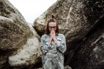 Mujer joven en gafas de pie junto a las piedras y rezando con los ojos cerrados . - foto de stock