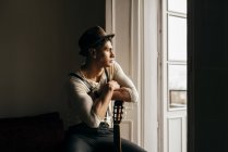 Homem atencioso de chapéu apoiado na guitarra e olhando para a janela — Fotografia de Stock