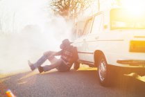 Homem com chapéu apoiado em carro quebrado emitindo fumaça na beira da estrada . — Fotografia de Stock