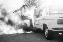 Man kicking broken vintage car emitting smoke on roadside. — Stock Photo