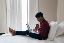 Чоловік з чашкою сидить на ліжку і переглядає ноутбук — стокове фото
