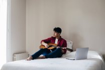 Человек с гитарой сидит на кровати и смотрит в сторону — стоковое фото