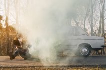Hombre con sombrero apoyado en coche vintage roto emitiendo humo estacionado en la carretera
. - foto de stock