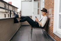 Hombre con ropa vintage sentado en el balcón y el teléfono inteligente de navegación - foto de stock
