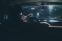Rear view of man in hat driving car with headlights on at night. — Stock Photo