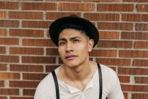 Stilvoller junger Mann mit Hut blickt auf Ziegelmauer in die Kamera. — Stockfoto