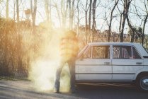 Vista trasera del hombre abriendo la capucha de fumar coche vintage roto en la naturaleza . - foto de stock