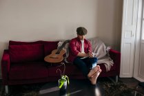 Jovem de boné sentado no sofá pela guitarra e tomando notas — Fotografia de Stock