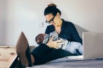 Femme souriante dans des lunettes assis sur le lit avec ordinateur portable et tenant son fils endormi . — Photo de stock