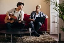 Молоді стильні чоловіки відпочивають на дивані з гітарою і ноутбуком — стокове фото