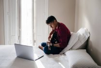 Uomo seduto con la chitarra e guardando il computer portatile sul letto — Foto stock