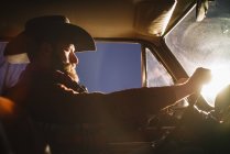 Vista lateral do homem em carro de condução chapéu ao pôr do sol luz — Fotografia de Stock