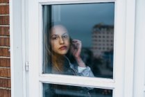 Jovem mulher tocando orelha e olhando para a câmera através da janela em casa . — Fotografia de Stock