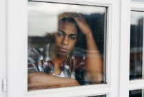 Thoughtful young man standing behind window and looking at camera. — Stock Photo