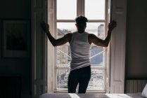 Back view of man standing at window and opening shutters in bedroom. — Stock Photo