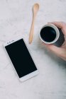 Обрізати чоловічу руку, тримаючи чорну каву в одноразовій чашці за допомогою смартфона — стокове фото