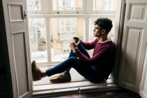 Man with cup sitting on window sill and looking away — Stock Photo