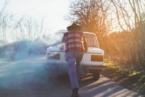 Back view of man in hat pushing broken vintage car in countryside. — Stock Photo
