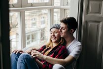 Joven hombre y mujer alegre sentado en el alféizar de la ventana y abrazándose en casa . - foto de stock