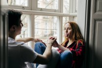 Young woman holding hands and showing tongue out to boyfriend on window sill at home. — Stock Photo