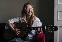 Mulher alegre em óculos sentado e tocando guitarra acústica em casa . — Fotografia de Stock