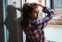 Young woman holding hair and looking back at camera by window. — Stock Photo