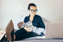 Brunette femme dans des lunettes tenant enfant endormi et ordinateur portable de navigation — Photo de stock